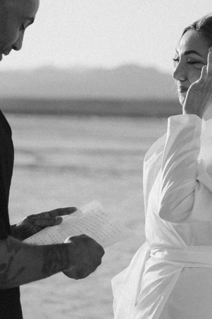 Black and white photo of a bride wiping her tears away as her groom reads his vows to her