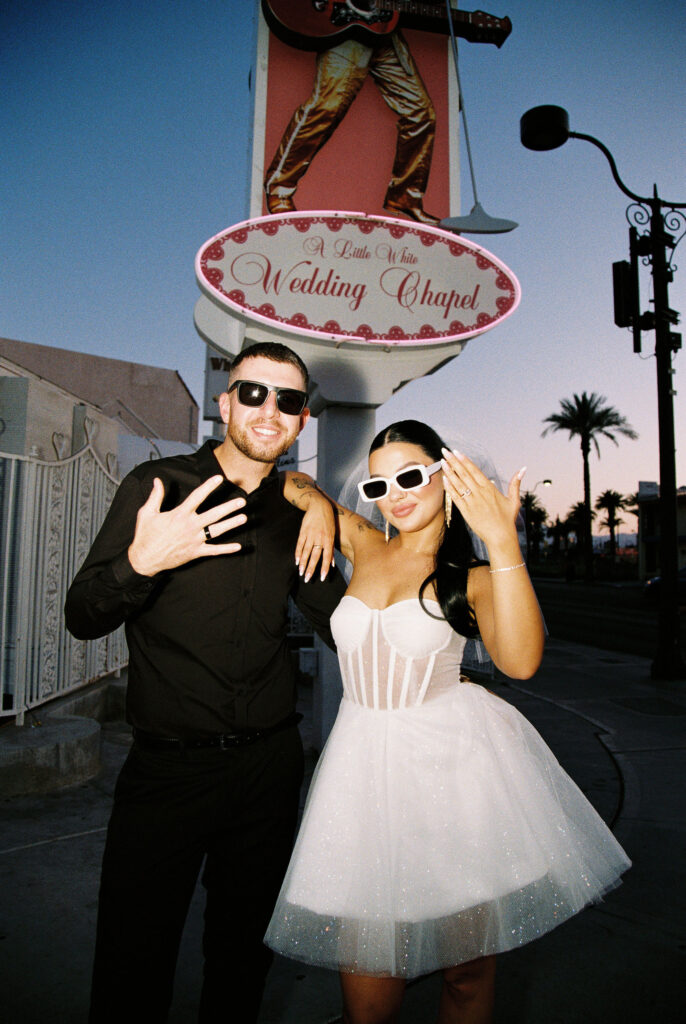 Bride and groom showing off their wedding rings in front of The Little White Wedding Chapel sign at night