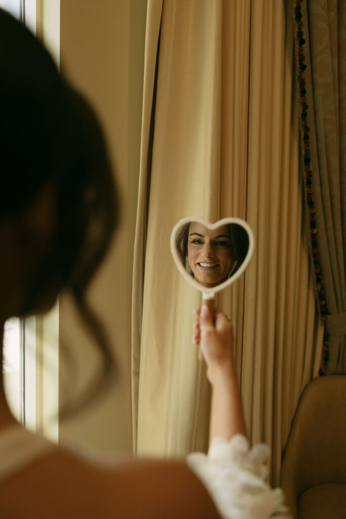 Bride looking at herself in a heart shaped mirror as she gets ready for her wedding day. 