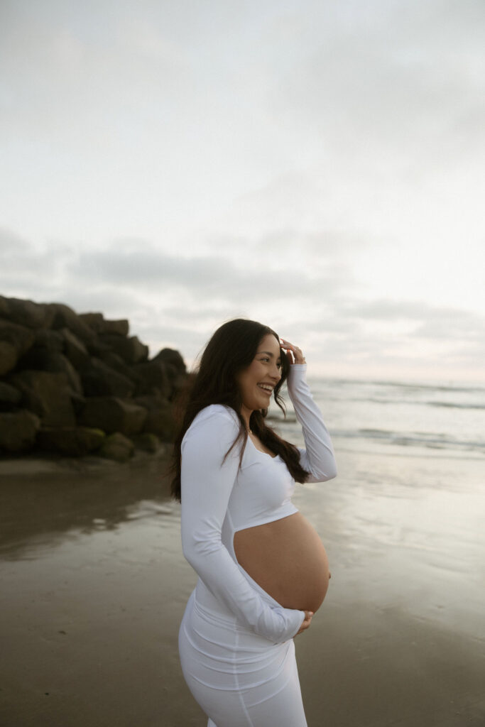 Woman laughing and holding her belly during her San Diego maternity photos on the beach. 