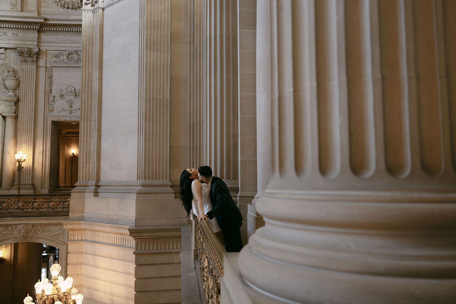 Man kissing his fiancés neck as they pose on the balcony for their San Francisco City Hall engagement photos