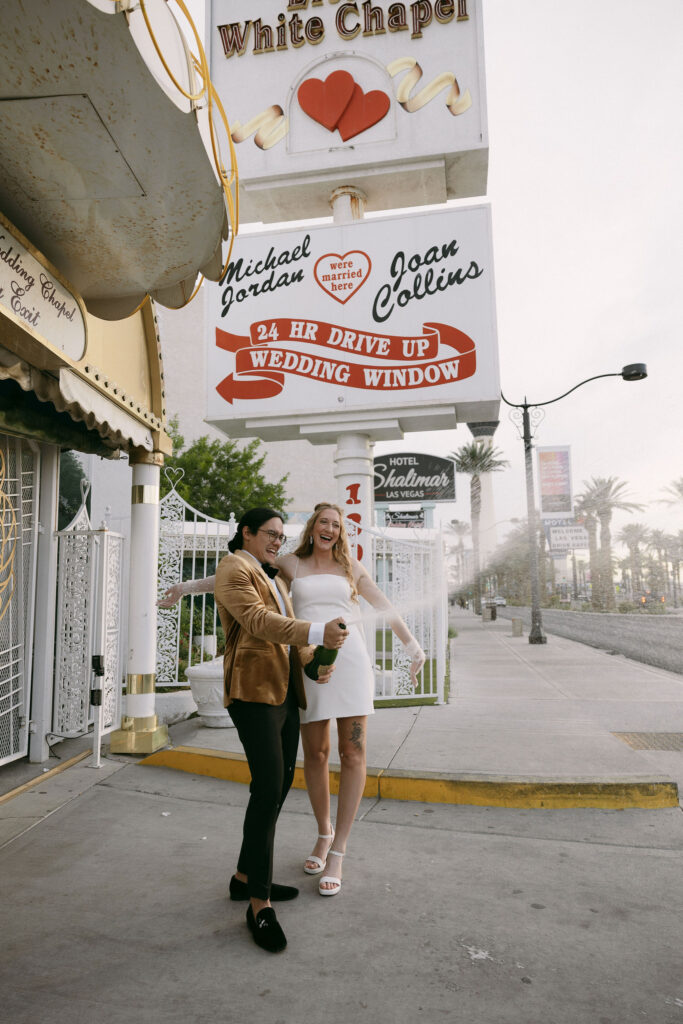 Bride and groom popping champagne in front of Little White Wedding Chapel