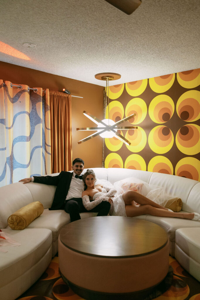 Bride and groom lounging on a white curved couch in a bold 70s-style room with orange and yellow retro wallpaper during their retro Las Vegas elopement.