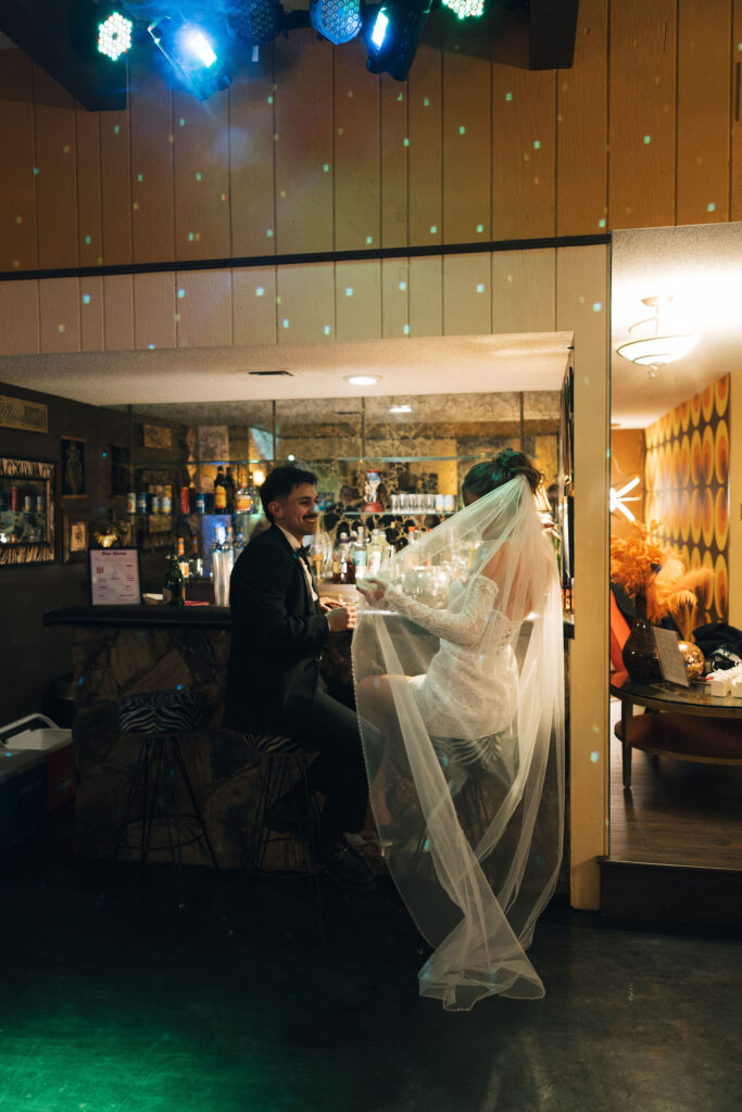 Bride and groom clinking glasses together at a vintage bar with disco lights shining during their retro Las Vegas elopement.