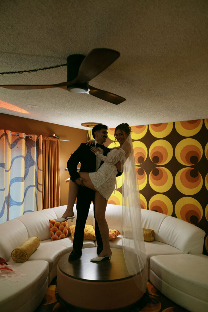 Groom lifting bride playfully on a coffee table in a vintage-inspired living room with groovy patterned walls at their retro Las Vegas elopement.