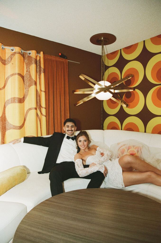 Bride and groom lounging on a white couch in a 70s retro Vegas home with bold patterned wallpaper.