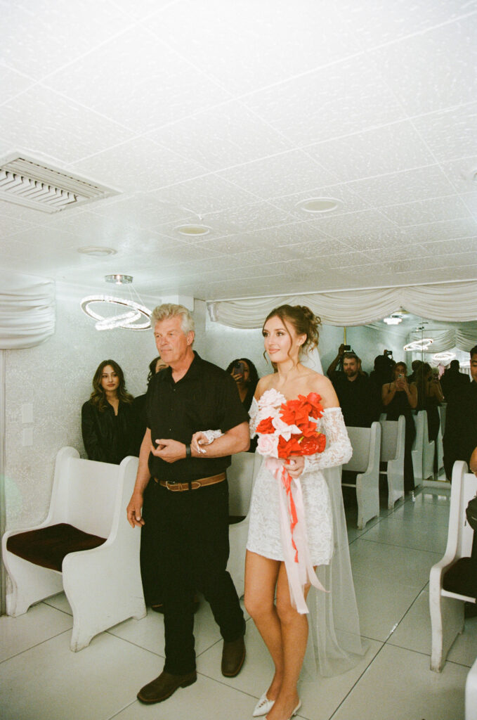 Bride walking down the aisle with her father inside The Little White Wedding Chapel on film