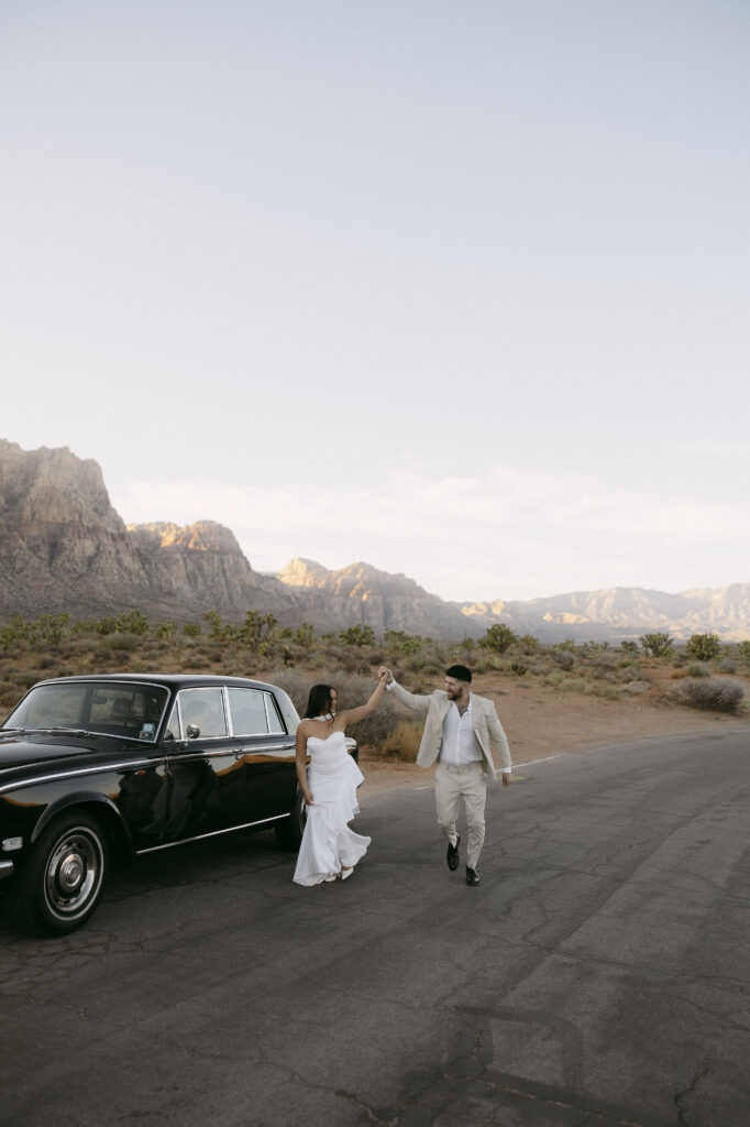 Couple holding hands beside a black classic car parked along the road at Red Rock Canyon.