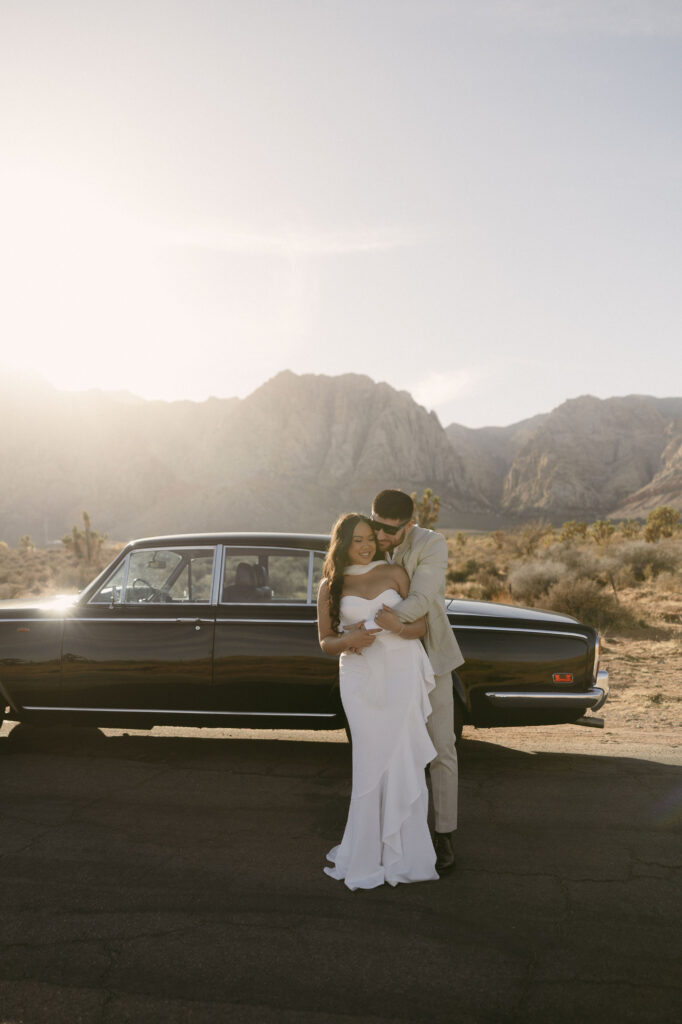 Couple embracing and laughing near their vintage car at Red Rock Canyon, surrounded by warm desert light.
