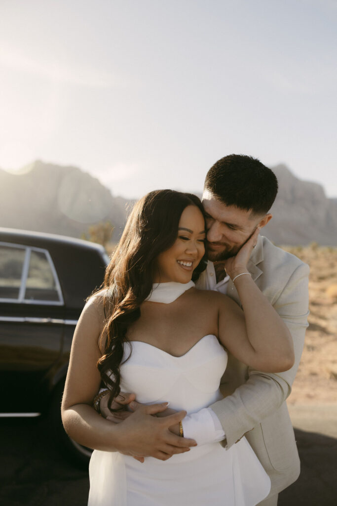 Couple embracing and laughing near their vintage car at Red Rock Canyon, surrounded by warm desert light.