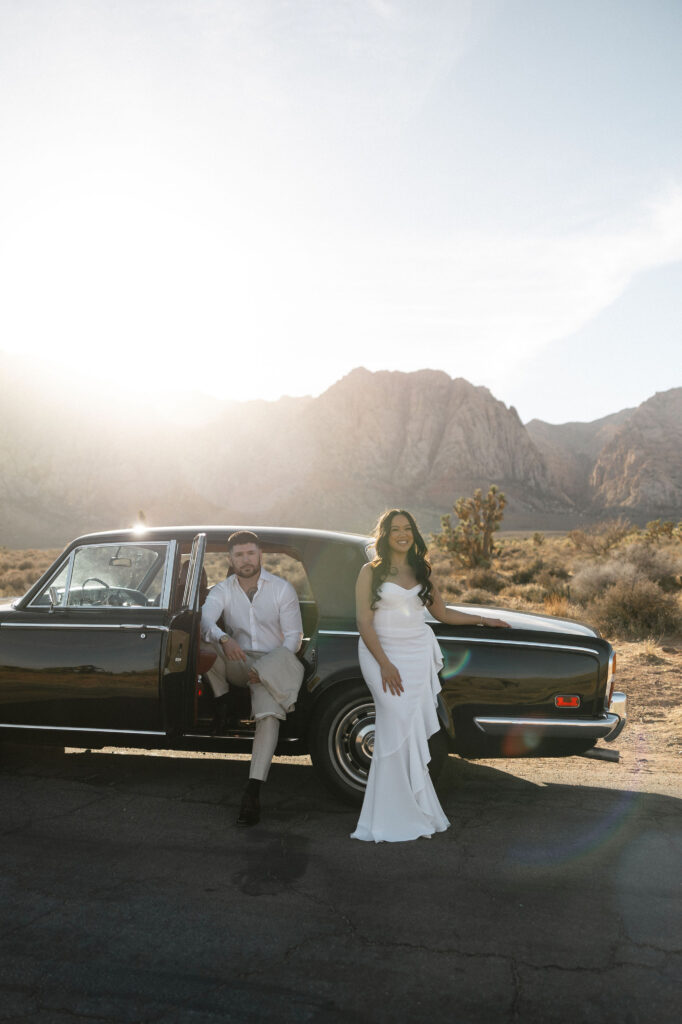 Couple standing beside a vintage black car with mountains in the background during Red Rock Canyon engagement photos.
