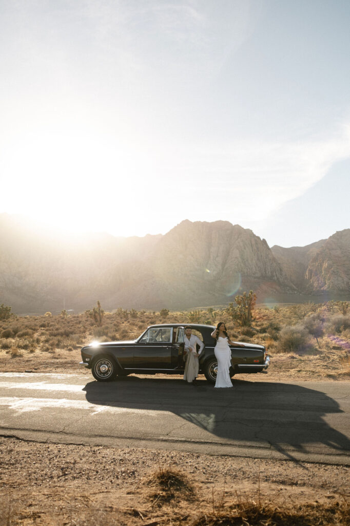 Couple standing beside a vintage black car with mountains in the background during Red Rock Canyon engagement photos.