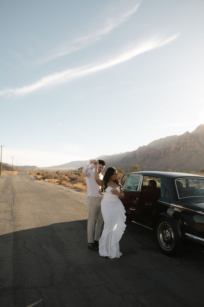 Couple twirling beside a vintage black car on a sunlit desert road at Red Rock Canyon.