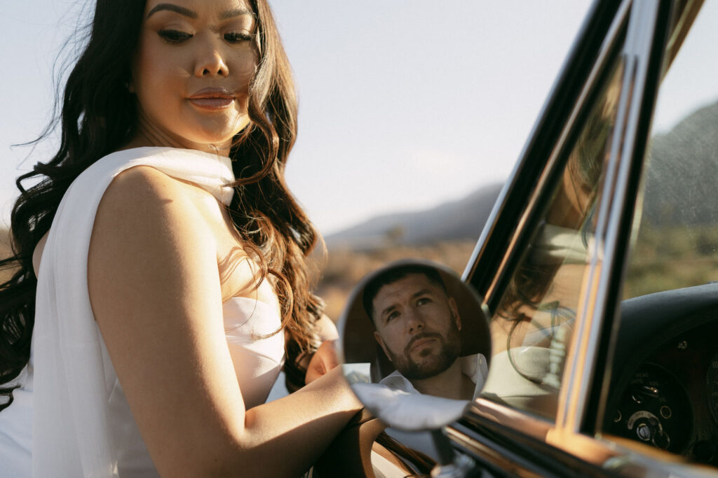 Reflection of a man’s face in a car mirror while his partner stands beside the car during Red Rock Canyon engagement photos.