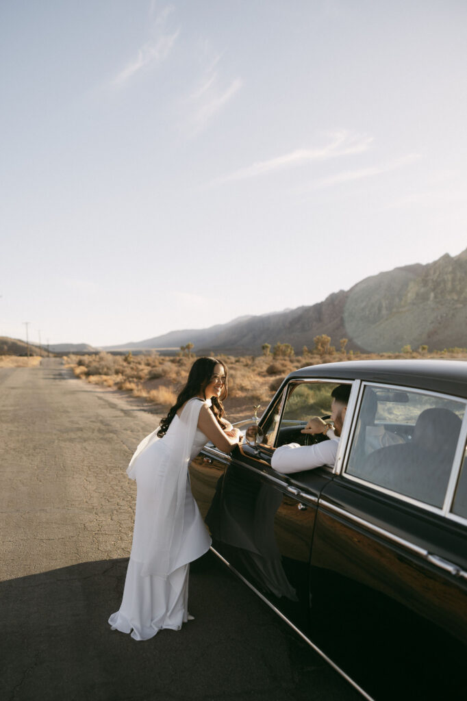 Woman leaning against a vintage car and smiling while talking to her partner on a desert road at Red Rock Canyon.