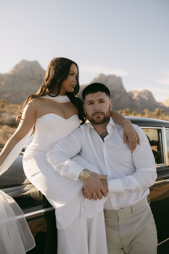 Couple sitting on the hood of a vintage black car surrounded by desert landscape during Las Vegas engagement photos.