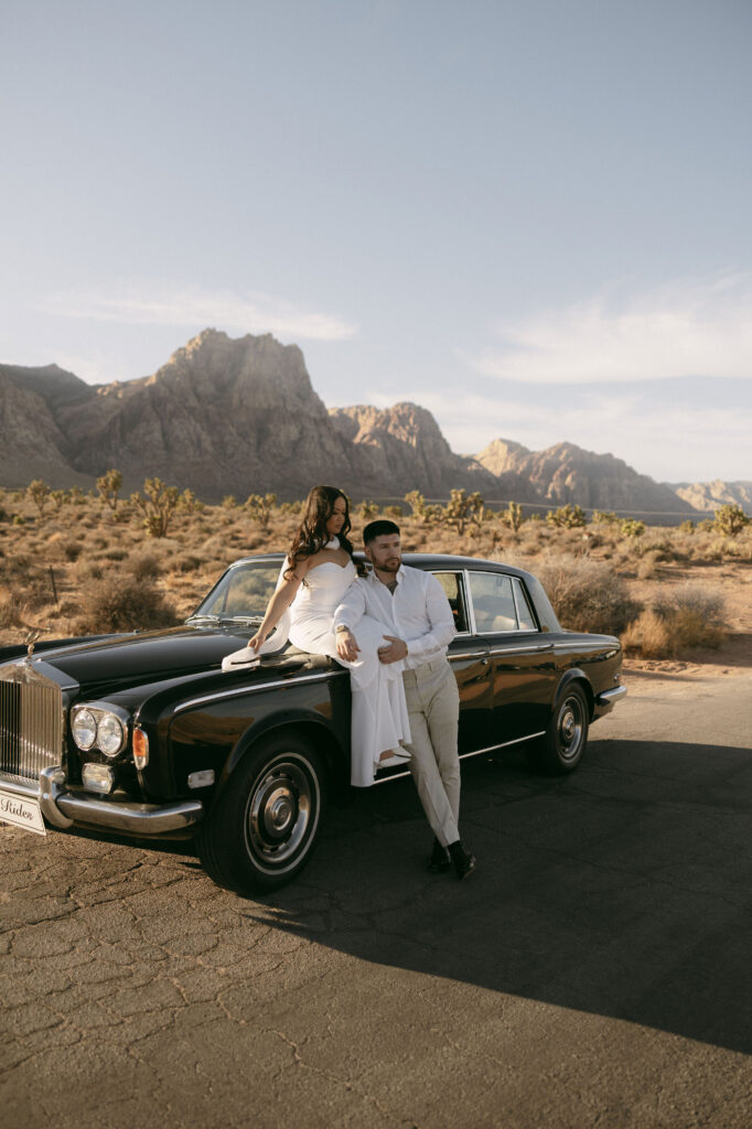 Couple posing together beside a black vintage car with desert mountains in the background during Red Rock Canyon engagement photos.