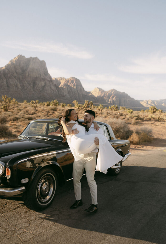Man lifting his fiance in front of their vintage Rolls-Royce with desert mountains behind them at Red Rock Canyon.