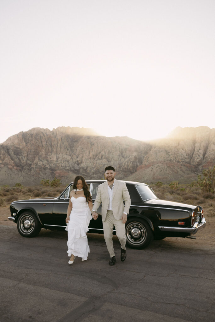 Couple walking hand-in-hand down the road at Red Rock Canyon, their classic car parked behind them.