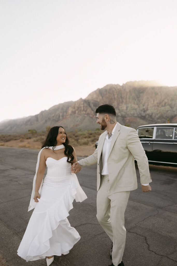 Couple walking hand-in-hand down the road at Red Rock Canyon, their classic car parked behind them.