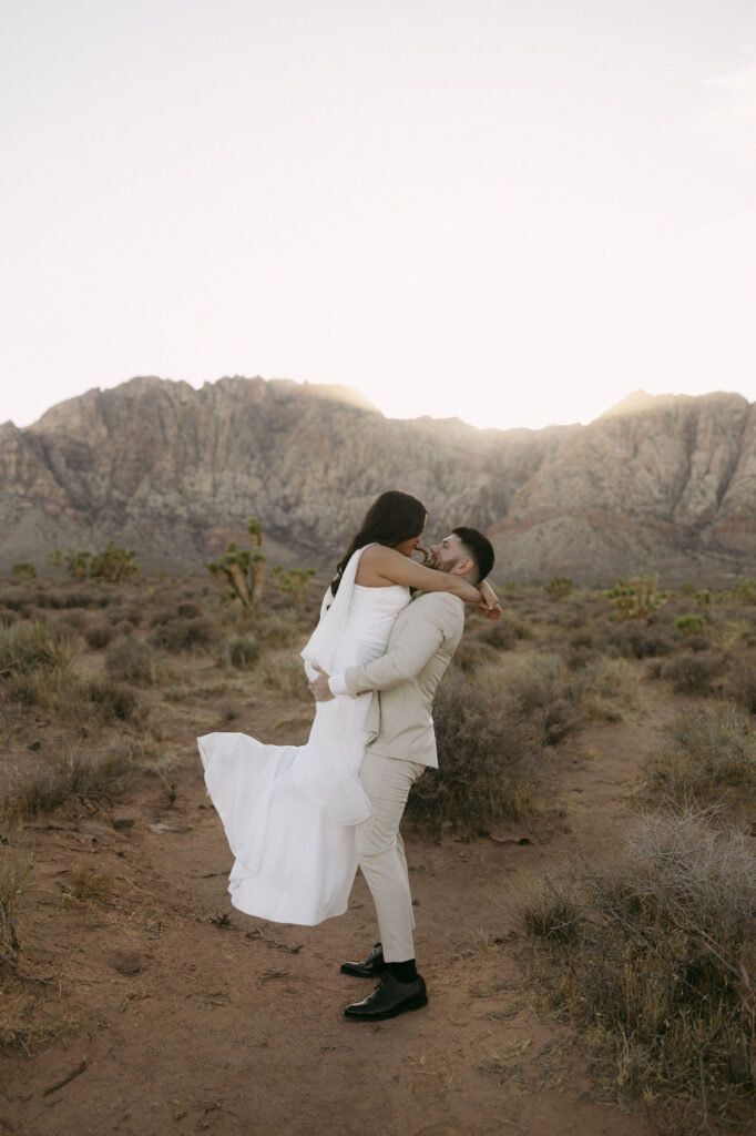 Man lifts his fiancé in the air while her white dress flows in the wind during their Red Rock Canyon engagement photos.