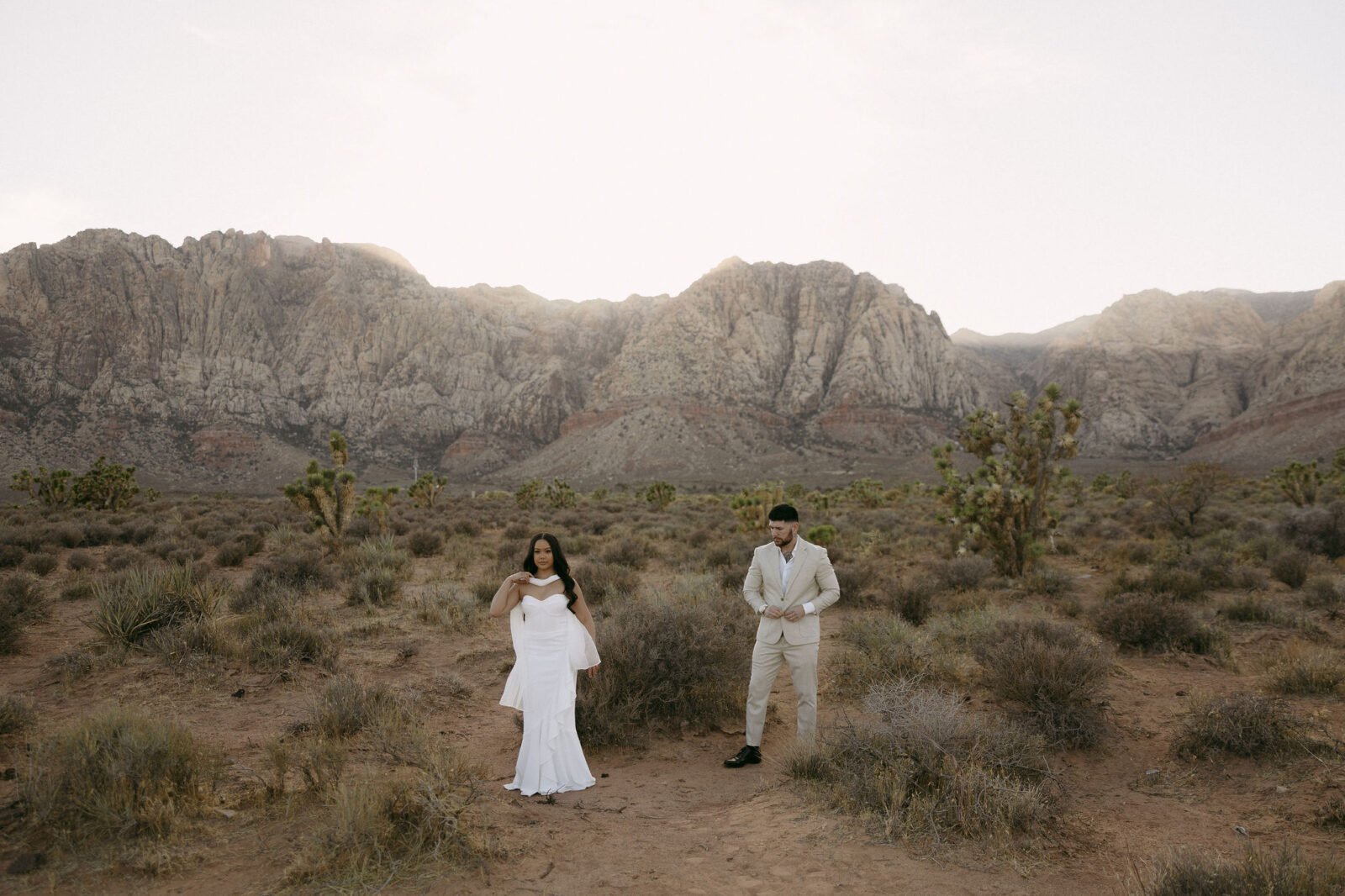 Couple standing apart in the desert landscape with mountains rising behind them during their Red Rock Canyon engagement session.