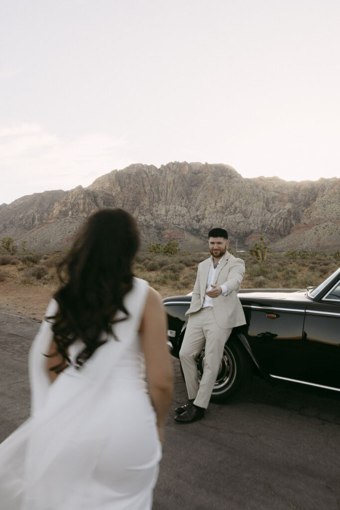 Woman walking toward her fiancé as he leans against a vintage car at Red Rock Canyon, mountains in the background.