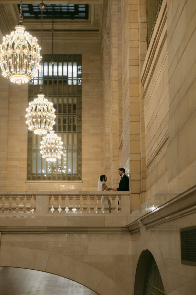 Couple standing together on the balcony inside Grand Central Station surrounded by glowing chandeliers.