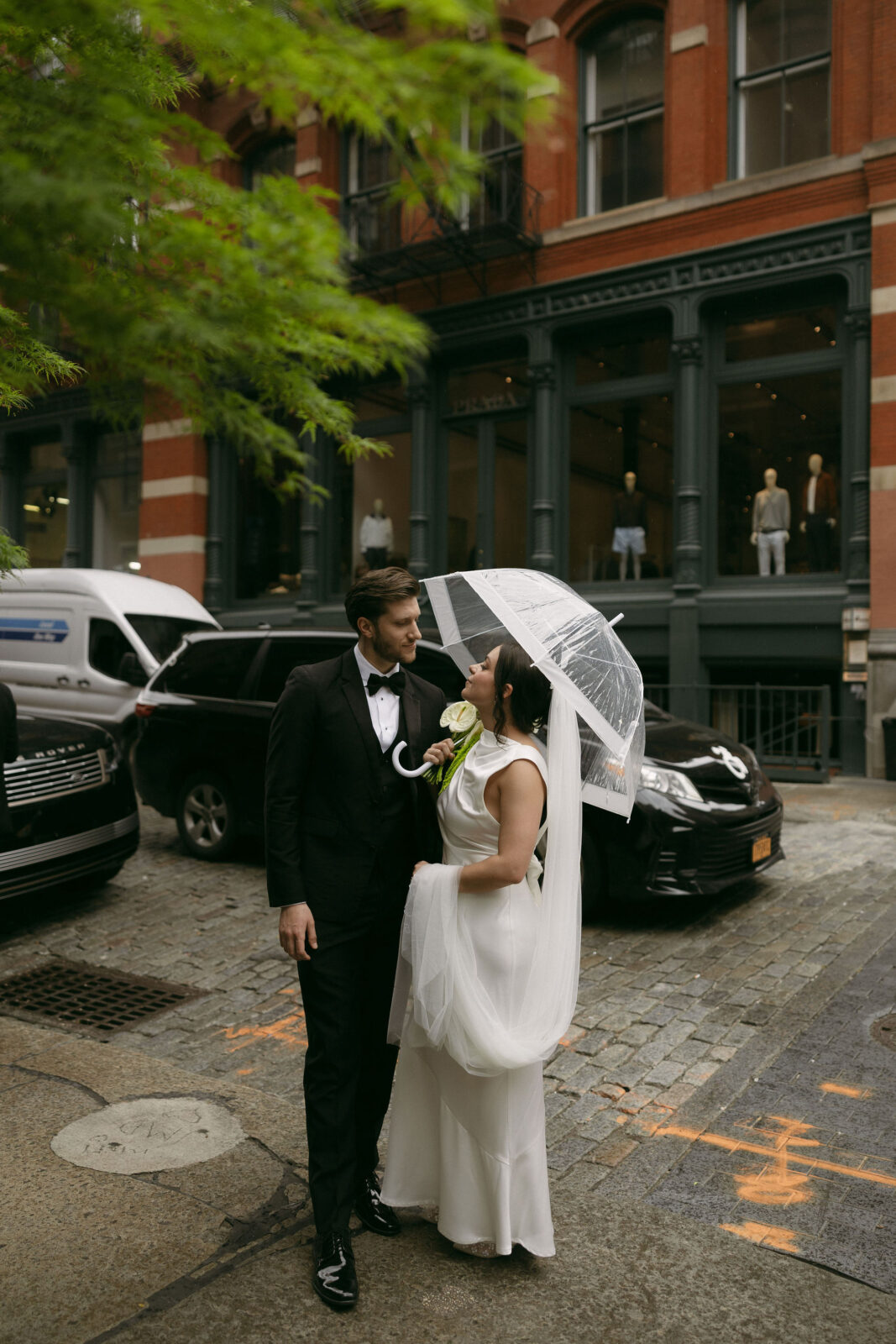 Bride and groom standing together under a clear umbrella on a cobblestone street after their New York City Hall elopement.